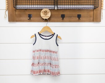 May include: A white sleeveless dress with a blue trim and a red and blue patterned bottom. The dress is hanging on a wooden hanger against a white wood background.
