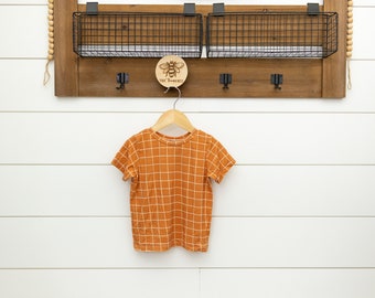 May include: A burnt orange t-shirt with a white checkered pattern hangs on a wooden hanger. The t-shirt is on a white wall with a wooden shelf above it. The shelf has two black wire baskets and a wooden sign with a bee design that says "The Honeybee".