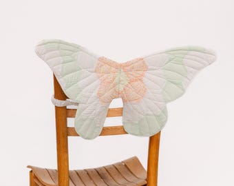 May include: A white and green butterfly-shaped chair back cushion with a pink and orange floral design. The cushion is attached to a wooden chair.