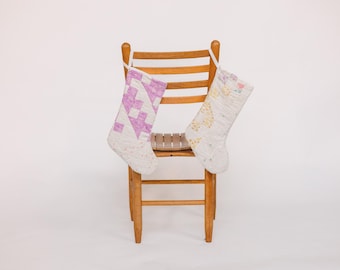 May include: Two fabric Christmas stockings hanging on a wooden chair. The stocking on the left is white with a pink and white geometric pattern. The stocking on the right is white with a yellow and white floral pattern.