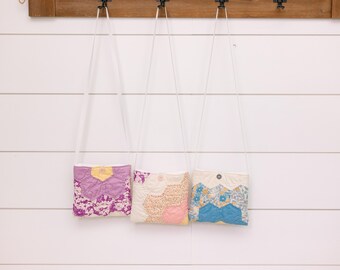 May include: Three small quilted bags hanging on hooks. The bags are made of different fabrics and have a patchwork design. The bag on the left is purple, the middle bag is pink and yellow, and the bag on the right is blue and white.