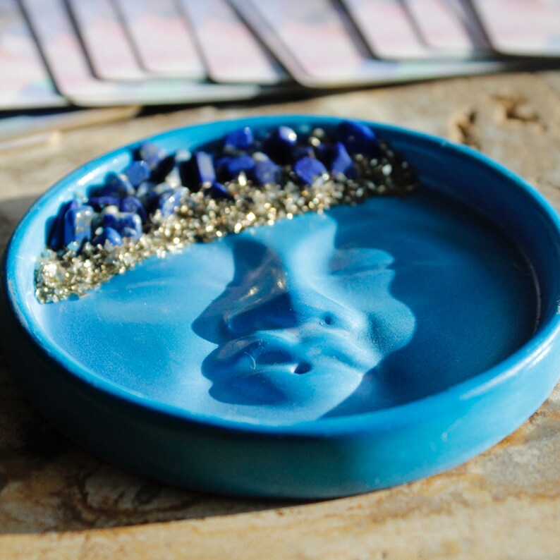 Pu&ograve; includere: Un piatto in ceramica blu con un viso scolpito sul fondo. Il piatto &egrave; decorato con un bordo di glitter blu e argento.