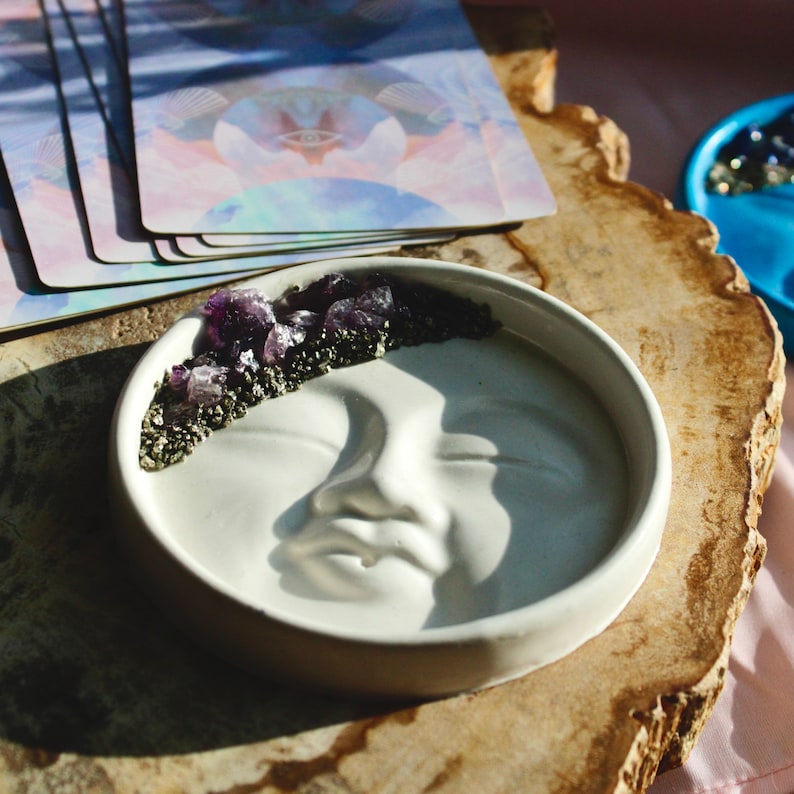 Pu&ograve; includere: Un piatto in ceramica bianca a forma di luna con un design a rilievo di un viso. Il piatto &egrave; decorato con un bordo di piccoli cristalli viola e dorati.