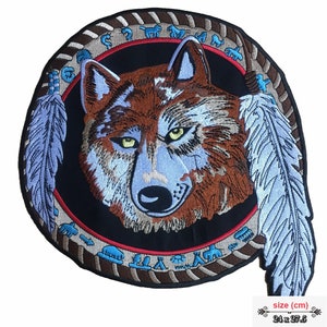 May include: Embroidered patch featuring a wolf's head with blue and brown fur, surrounded by a brown and tan circular frame with Native American symbols. Two blue feathers extend from the frame. The patch measures 24 x 27.5 cm.