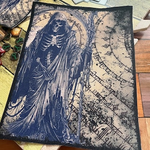 May include: A blue and white embroidered patch featuring a skeletal figure in a hooded robe holding a staff. The figure is standing in front of a circular design with intricate patterns and text.