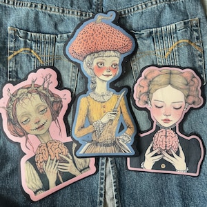 May include: Three embroidered iron-on patches featuring illustrations of women with brains. One woman wears a red mushroom hat and holds a knife, another wears a crown of thorns and holds a brain, and the third has her eyes closed and holds a brain.