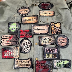 May include: A collection of distressed-style patches with various phrases and words related to mental health, such as "Asylum," "Delusion," and "Inner Noise." The patches are in various shapes and sizes, with a vintage aesthetic, and are affixed to a green denim jacket.
