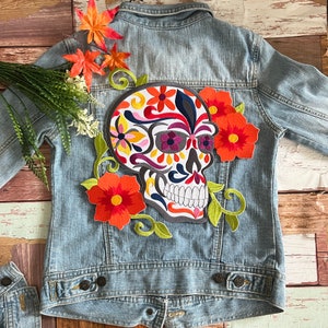 May include: A blue denim jacket with an embroidered sugar skull patch. The skull is decorated with colourful flowers and swirls.