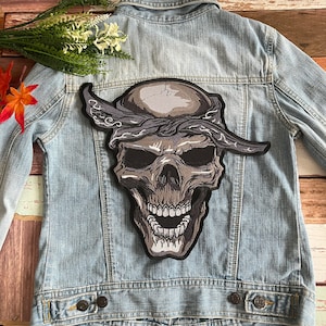 Könnte beinhalten: Ein schwarzer und grauer Schädel mit einem Bandana auf dem Kopf. Der Schädel ist auf eine Jeansjacke gestickt.