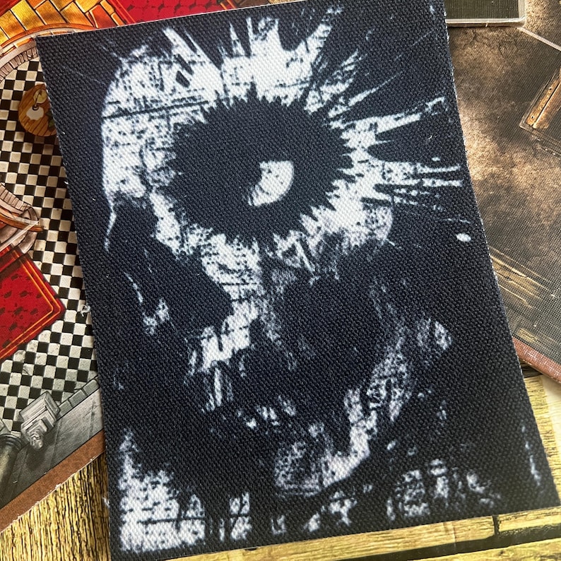 May include: Black and white embroidered patch featuring a skull design. The skull has a large eye and a distressed, textured appearance.