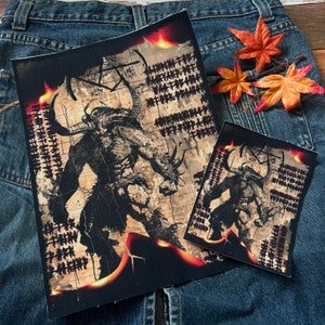 Peut inclure: Deux patchs en tissu noir avec une illustration brune et noire d'une créature cornue avec des flammes sur un fond de parchemin. Les patchs sont sur un fond de denim bleu.