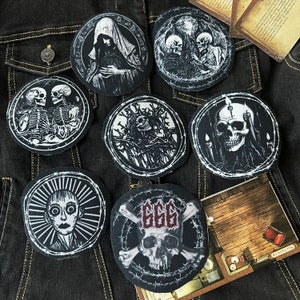 Könnte beinhalten: Sieben gestickte Patches in Schwarz und Weiß mit verschiedenen Designs, darunter Schädel, Skelette und eine Kapuzenfigur. Die Patches sind auf eine dunkelblaue Jeansjacke genäht.