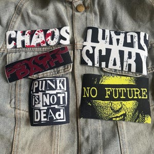 Op de afbeelding: Een spijkerjack met vijf strijklabels. De labels zijn zwart-wit met rode en gele accenten. De labels tonen de tekst "Chaos", "Psycho", "Punk is Not Dead", "Lousy Scary" en "No Future".