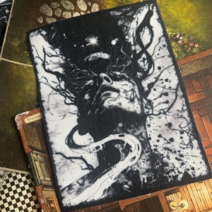 May include: Black and white embroidered patch featuring a stylised image of a face with a swirling, tree-like design. The background is black with white splatter paint.