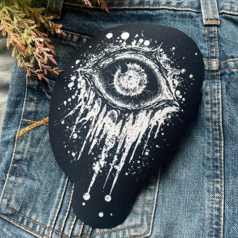 May include: Black and white embroidered patch with a large eye design. The eye is surrounded by white paint splatters.