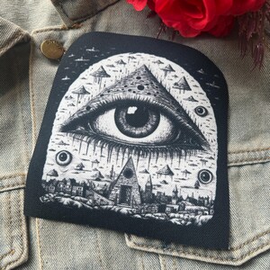 May include: Black and white embroidered patch with a large eye inside a triangle, surrounded by smaller triangles and circles. The eye is looking directly at the viewer.