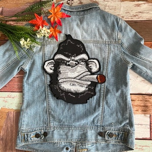 Könnte beinhalten: Eine blaue Jeansjacke mit einem schwarzen und weißen gestickten Aufnäher eines Affen, der eine Zigarette raucht. Der Aufnäher lautet "13th Titan".