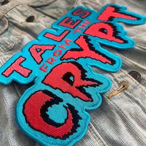 Könnte beinhalten: Eine hellblaue Jeansjacke mit einem großen gestickten Aufnäher auf der Rückseite. Der Aufnäher zeigt den Text TALES FROM THE CRYPT in leuchtend roten Buchstaben mit schwarzen Umrandungen auf einem leuchtend cyanblauen Hintergrund.