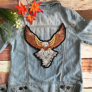 Könnte beinhalten: Ein gestickter Aufnäher mit einem braunen und goldenen Adler, der seine Flügel weit ausgebreitet hat und einen detaillierten Motorradmotor in seinen Krallen hält. Der Aufnäher ist zum Aufbügeln geeignet und perfekt, um Jeansjacken, Westen oder Taschen einen einzigartigen Touch zu verleihen.