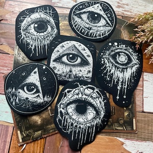 May include: Six black and white embroidered patches with an all-seeing eye design. The patches feature a variety of designs, including a pyramid, a clock, and a dripping eye.