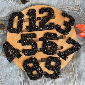 Könnte beinhalten: Ein Satz von zehn schwarzen Strasszahlen von 0 bis 9, die in einem Kreis auf einem Holzgrund angeordnet sind.