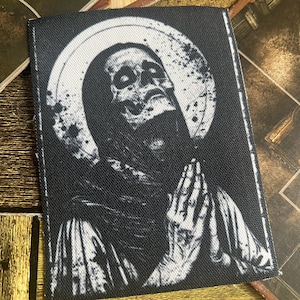 May include: Black and white embroidered patch depicting a skeletal figure with its hands clasped in prayer, surrounded by a halo. The figure is wearing a hooded robe and has a distressed, aged appearance.