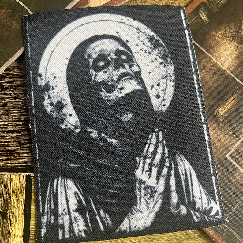 May include: Black and white embroidered patch depicting a skeletal figure with its hands clasped in prayer, surrounded by a halo. The figure is wearing a hooded robe and has a distressed, aged appearance.