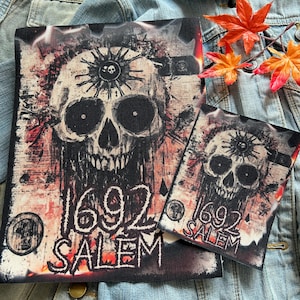 May include: Two iron-on patches with a skull and crossbones design. The larger patch features a skull with a third eye and the text "1692 SALEM". The smaller patch features a similar skull with the text "1692 SALEM".