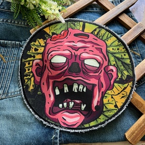May include: A round, colorful patch with a cartoon zombie face. The zombie has pink skin, a wide open mouth with teeth, and white eyes. The background is a green and yellow leafy pattern.