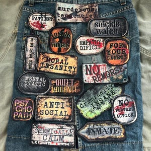 Könnte beinhalten: Ein Jeanskleidungsstück, das mit verschiedenen rechteckigen und ovalen Aufnähern verziert ist. Die Aufnäher zeigen abgenutzte Texte wie "Patient", "Inside Voices", "Moral Insanity" und "Criminally Insane". Die Farbpalette umfasst Rot, Schwarz, Weiß und Brauntöne.