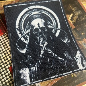 May include: Black and white embroidered patch featuring a skeletal figure with a large head and horns, surrounded by a circular design.
