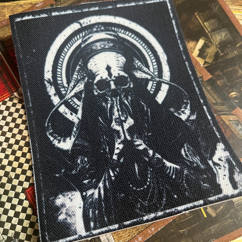May include: Black and white embroidered patch featuring a skeletal figure with a large head and horns, surrounded by a circular design.