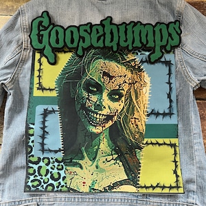 May include: A denim jacket featuring a green "Goosebumps" patch and a colourful graphic of a zombie woman with a stitched-up face. The graphic is surrounded by a black barbed wire frame.