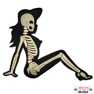 Può includere: Uno scheletro bianco di una donna seduta con le gambe incrociate, con un cappello nero. Lo scheletro è ricamato su una toppa di tessuto nero. La toppa misura 25 x 21,5 cm.