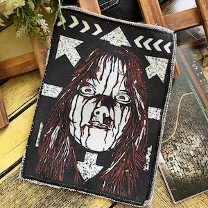 May include: A rectangular fabric patch featuring a black and white graphic illustration of a face with long hair and dripping paint effects. The design includes white arrows pointing in various directions against a solid black background.