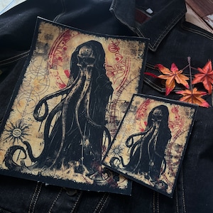 Può includere: Due toppe ricamate in bianco e nero che raffigurano una creatura tentacolare con un viso a forma di teschio. Le toppe sono su uno sfondo di jeans con un design rosso e dorato.