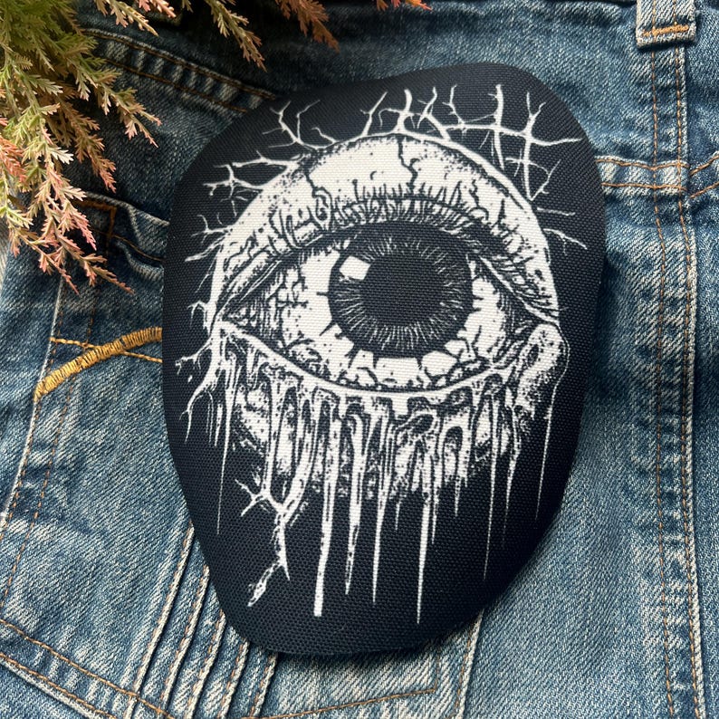 May include: Black and white embroidered patch of a large eye with dripping liquid, on a denim background.