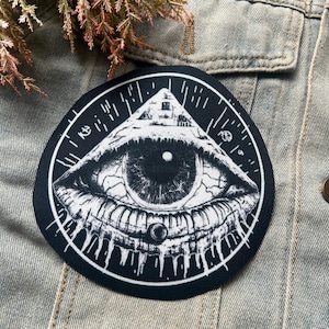 May include: Black and white embroidered patch with an eye inside a triangle. The eye is surrounded by a circle with lines radiating outward.