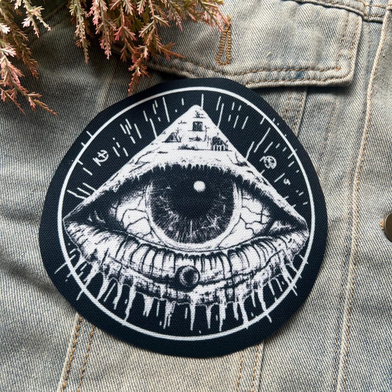 May include: Black and white embroidered patch with an eye inside a triangle. The eye is surrounded by a circle with lines radiating outward.