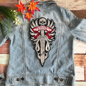 Könnte beinhalten: Ein gestickter Patch in Schwarzweiß mit einem Totenkopf, Flügeln, einem Schwert und dem Text "Choppers" in Rot. Der Patch ist auf eine blaue Jeansjacke genäht.