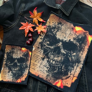 May include: Two black and white skull patches with a distressed, vintage look. The patches are on a denim jacket with a brown and orange background. The patches have a fiery orange border.