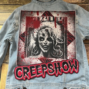 May include: A blue denim jacket with a red and black patch featuring a woman's face with a bloody mouth and the text "CREEP SHOW".