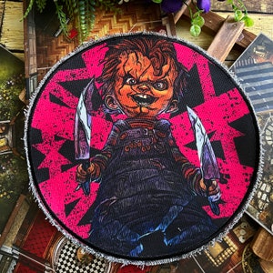 Può includere: Un cerotto rotondo in tessuto rosa e nero con un'immagine di un bambino dei cartoni animati che tiene due coltelli. Il bambino indossa una salopette blu e rossa e ha un sorriso malizioso. Il testo "Good Guy" è visibile sulla salopette.