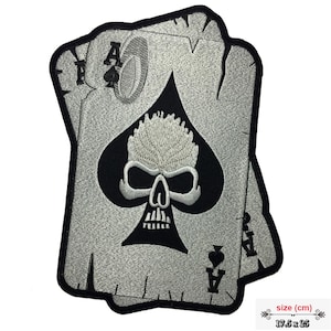 Könnte beinhalten: Gestickter Aufnäher mit einem Totenkopf-Design in einem schwarzen Pik-Form, vor einem verwitterten grauen Hintergrund. Der Aufnäher enthält die Symbole des Pik-Ass. Größe: ca. 17,5 x 25 cm.