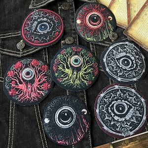 May include: Six embroidered patches with a black background and a white eye in the centre. The patches have different designs, including a tree, a sun, and a barbed wire fence. The patches are all about 7.5 cm in diameter.
