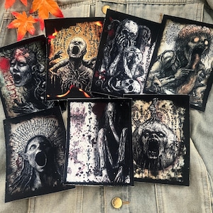 May include: A collection of seven black patches featuring detailed, grayscale illustrations of grotesque figures with open mouths and distorted features. The patches are arranged on a denim jacket, with autumn leaves in the background.