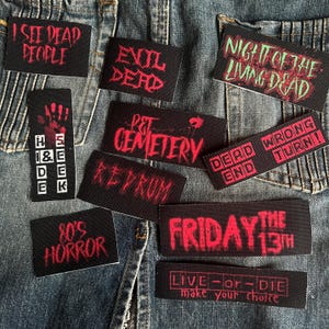 Könnte beinhalten: Eine Sammlung von neun schwarzen und roten gestickten Patches mit Horrorfilm-Motiven. Die Patches zeigen Titel wie "Evil Dead", "Pet Sematary", "Freitag der 13.", "Die Nacht der lebenden Toten" und "Ich sehe tote Menschen".