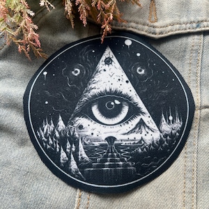 May include: Black and white embroidered patch with an all-seeing eye inside a triangle. The eye is surrounded by a dark forest and stars.
