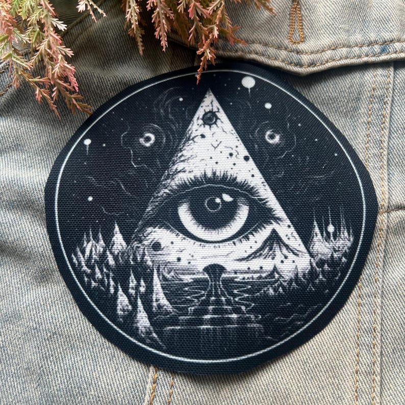 May include: Black and white embroidered patch with an all-seeing eye inside a triangle. The eye is surrounded by a dark forest and stars.