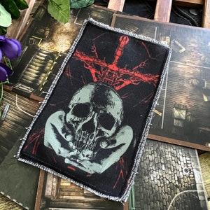 May include: A black and red embroidered patch featuring a skull in the centre, surrounded by two hands. The skull has a detailed design and is surrounded by a red, branching pattern.